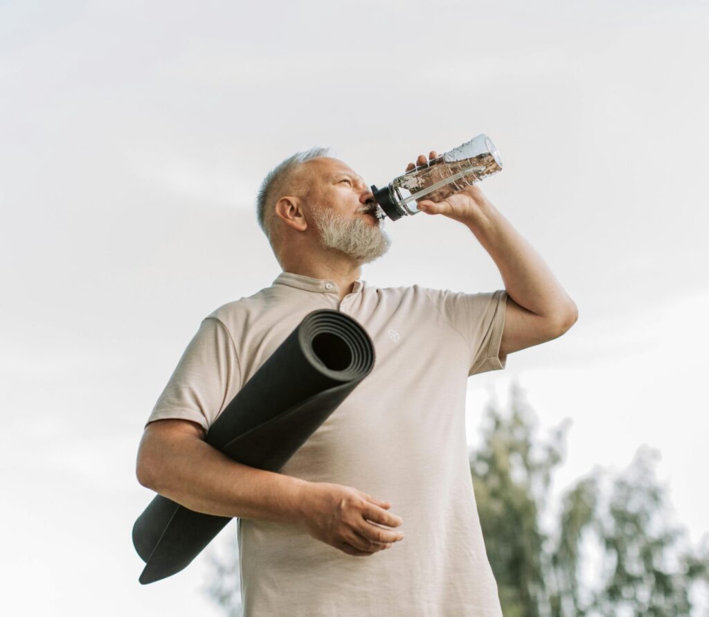 Elderly man drinking water while holding a yoga mat outdoors, promoting fitness and hydration.
